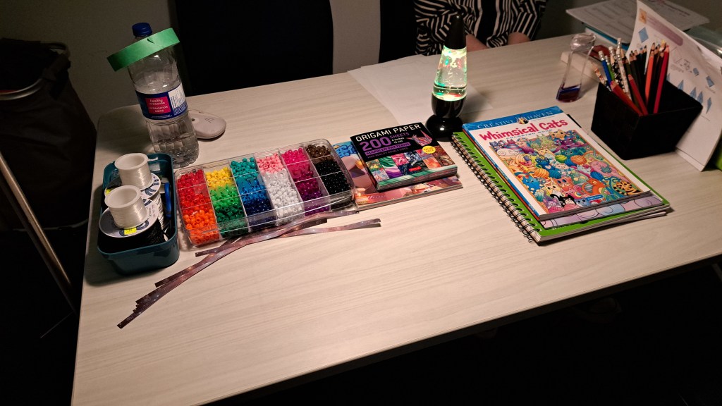 A variety of crafting materials including colourful beading supplies, colouring and sketch books, a tornado lamp, and origami paper on top of a table.